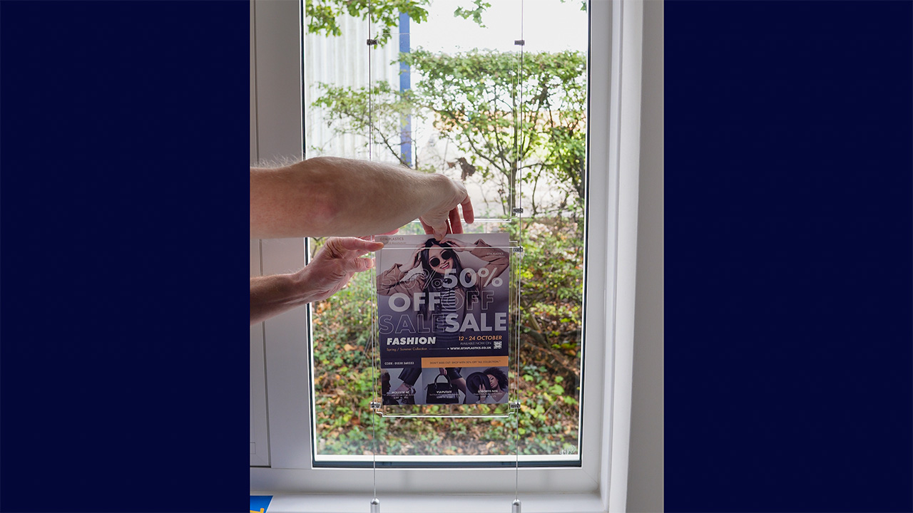 A person's hand inserting a poster into an acrylic poster holder that's suspended between two cables.